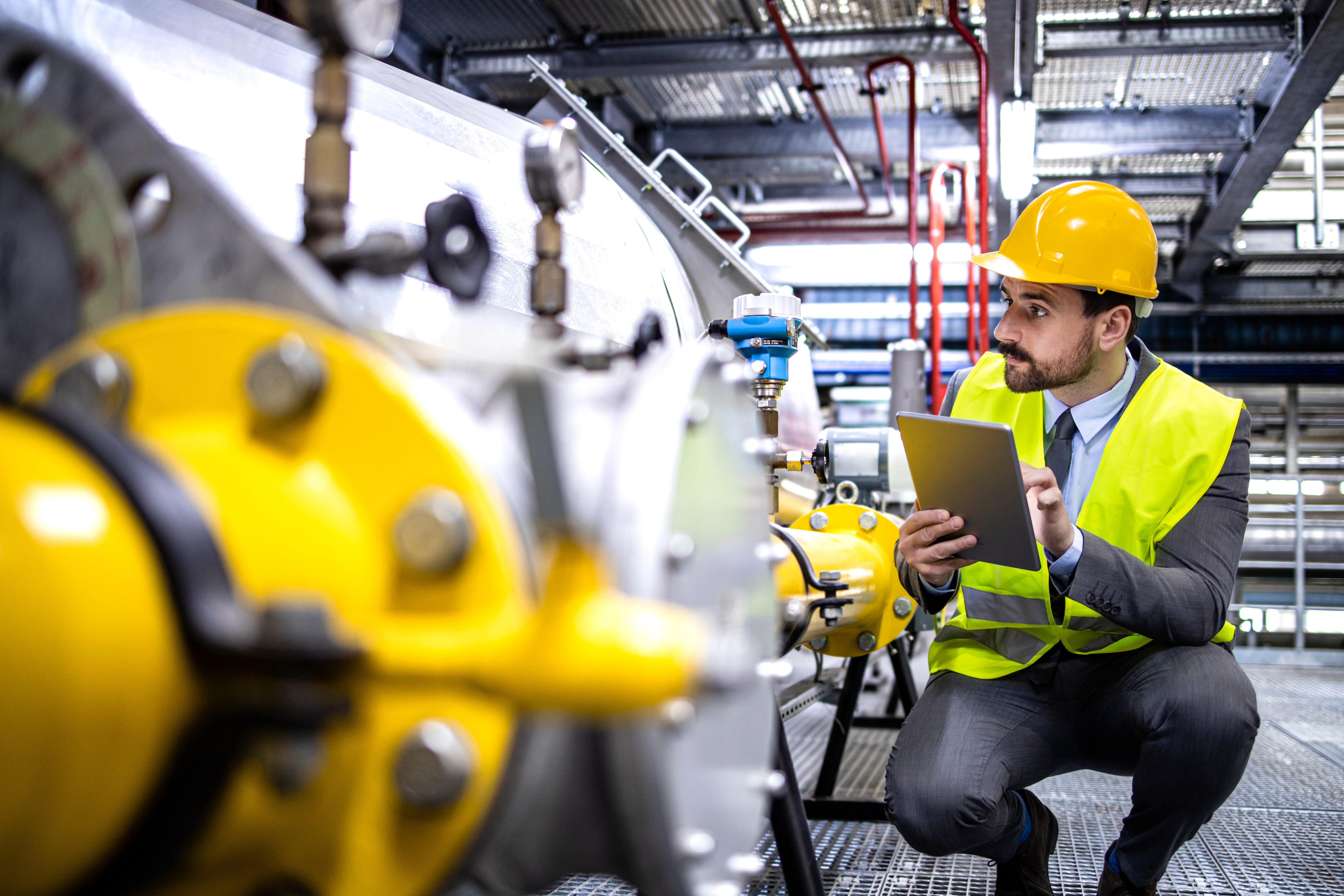 A technician wearing a helmet and safety vest monitors an industrial plant with a tablet in his hand.