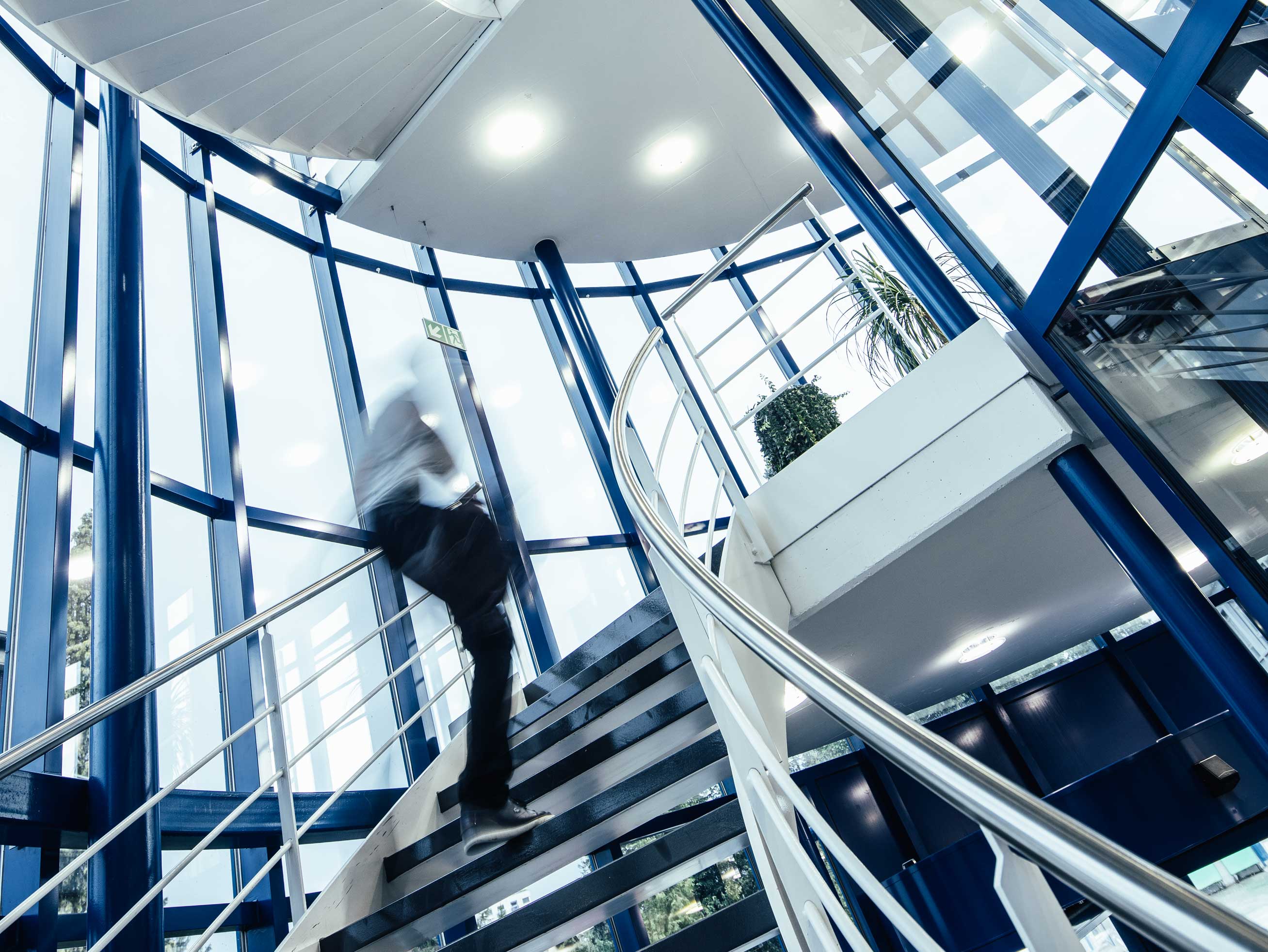 A person walks up a spiral staircase in a modern building with a glass facade and glass roof.