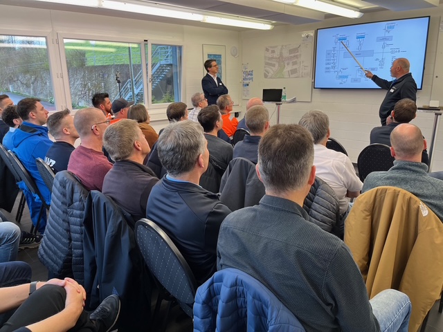 The speaker presents a technical diagram on a screen to a seated audience in a training room.