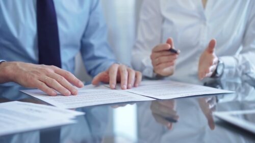 Two people in business attire are discussing a document on a glass table; one hand is holding a pen.